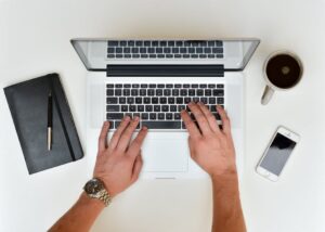 Hands typing on laptop with smart devices sitting on desktop signifying many ways employers can use Fingercheck without a physical time clock.