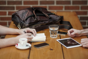 employees work outside at wood table, coffee, notepads, tablet, water.