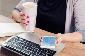 Employee looking at Fingercheck mobile app on smartphone at desk with coffee and laptop.