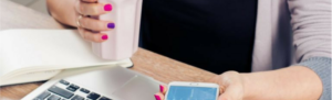Employee looking at Fingercheck mobile app on smartphone at desk with coffee and laptop.