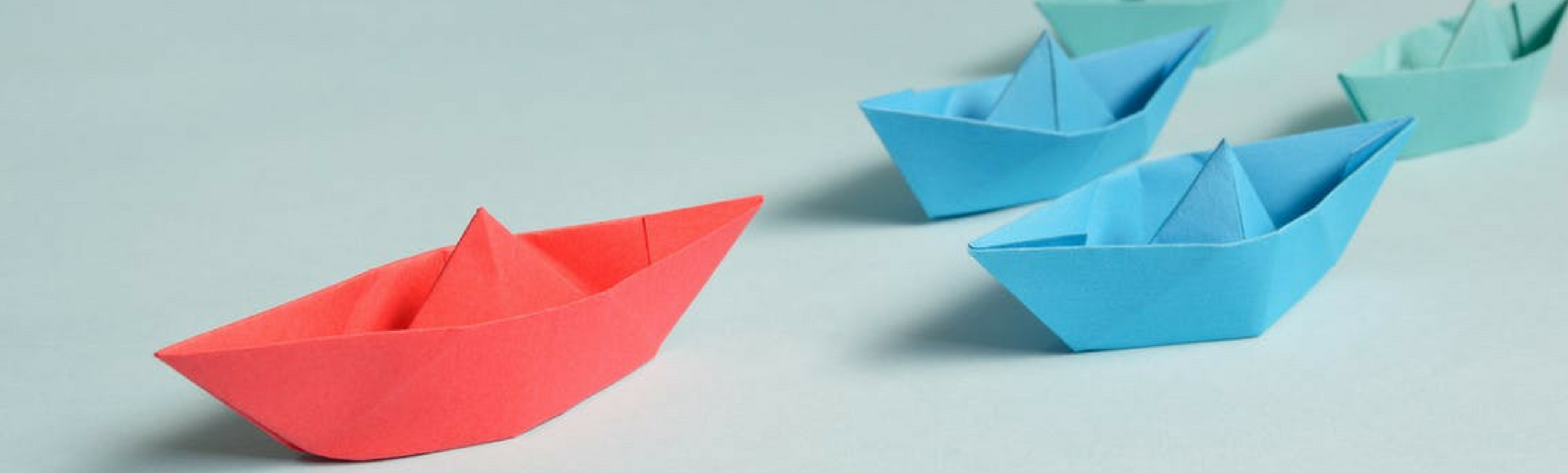 Small red oragami boat leads the way for blue boats and green boats behind it, reflecting show-don't-tell leadership tactics.