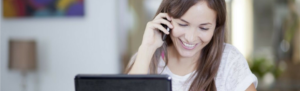 Employee working remotely from home holding smartphone to ear and talking while multi-tasking on laptop.
