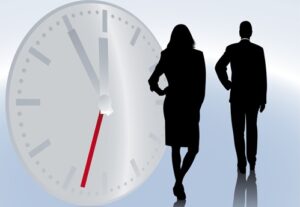Shadowy figures of businessman businesswoman standing in front of large time clock signifying time worked whether it's part-time or full-time, and the need for staffing solutions.
