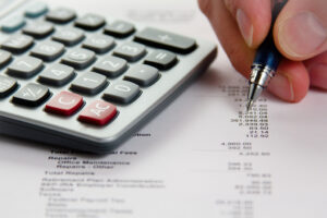 Close-up shot of payroll manager's hands doing a payroll reconciliation by hand with pen, calculator, referencing sheet of figures.