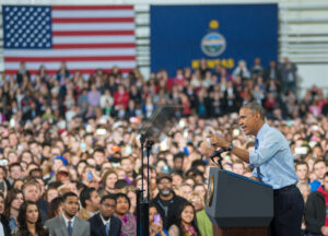 President Obama stands at podium with large crowds and U.S. flag backdrop announcing landmark policy extending overtime pay to 4.2 million salaried workers.