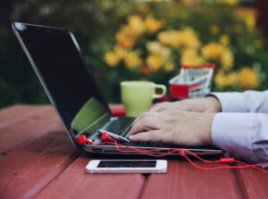 Relaxed professional on laptop in backyard typing with smartphone and coffee cup alongside, using web-based business solution to manage tasks paperlessly.