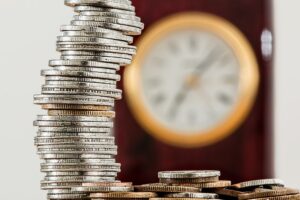 Brass clock with white face in rich mahogany casing and pile of coin currency signifies the financial benefits of automating time and attendance.