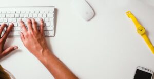 Hands typing white keyboard, mouse, yellow watch, using new feature from Fingercheck to reprint checks directly from employees profiles.