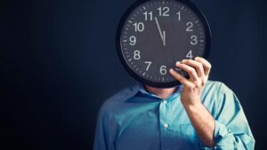 Business man holds time clock over face to represent an overnight shift and Fingercheck's new time card icon.