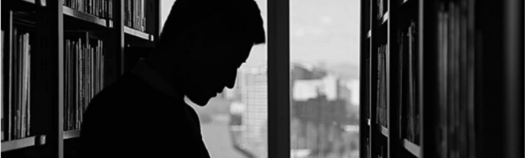 Man shrouded in shadows in the stacks of a library with window in background, solemn position, represents employers reading labor laws or else be penalized.