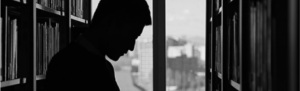 Man shrouded in shadows in the stacks of a library with window in background, solemn position, represents employers reading labor laws or else be penalized.